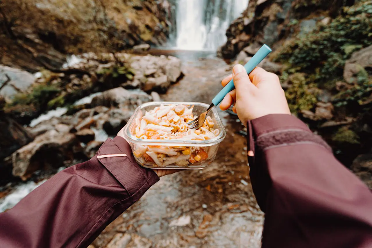 backpacker-eating-food-by-waterfall Backpacker eating pasta at camp near waterfall — adjusting food style and calorie planning on trail.