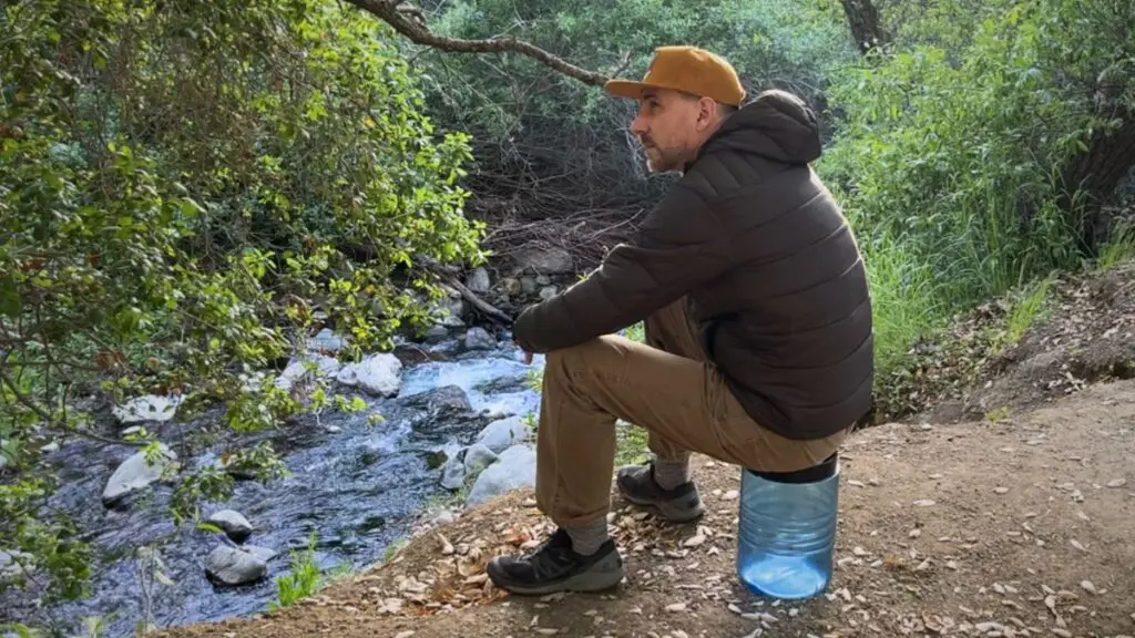 backpacker-sitting-on-bear-canister-river Backpacker in a puffy jacket sitting on a bear canister beside a creek