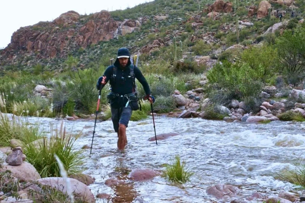 backpacker-crossing-river-trekking-poles Man with backpack and trekking poles wading barefoot through rocky desert stream with saguaro cacti and red rock hills in background