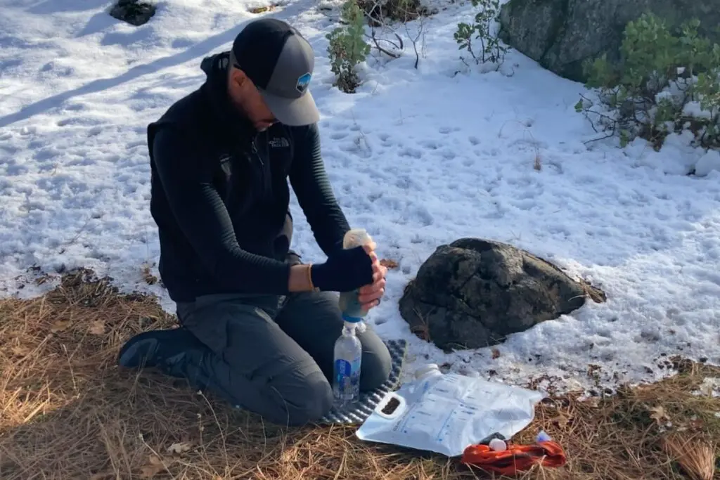 backpacker-filtering-water-in-the-snow Hiker kneeling in the snow filling a gravity water filter bag, with a clear bottle and filter setup laid out on the ground.