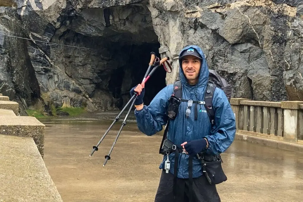 backpacker-holding-trekking-poles Smiling hiker in blue rain jacket holding trekking poles in front of a flooded rock tunnel on a rainy day