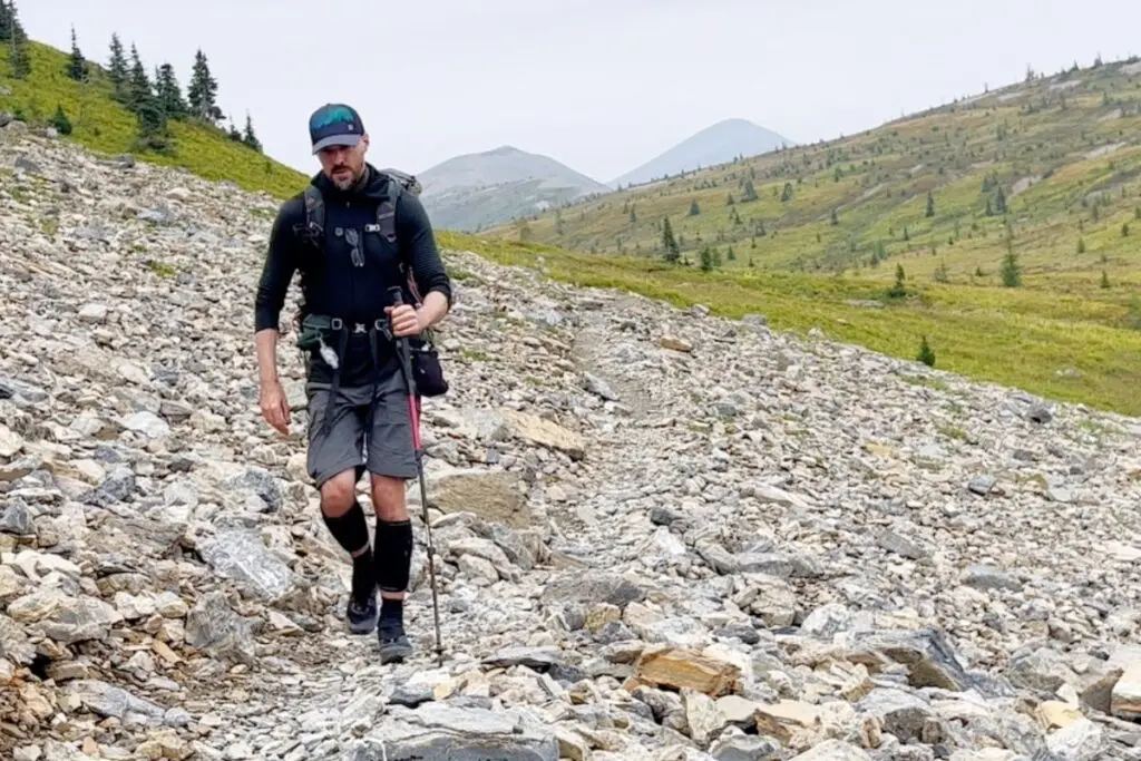 backpacker-trekking-poles-rough-terrain Man hiking across rocky mountain scree field with trekking pole and backpack, wearing knee brace