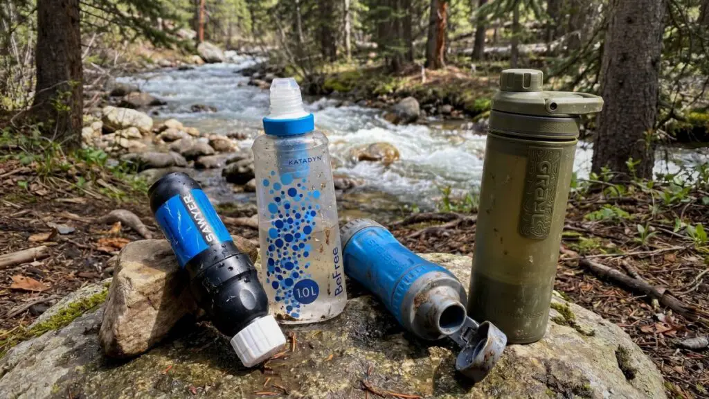 backpacking-water-filters-by-river Four backpacking water filters lined up on a rock beside a flowing creek: a Sawyer squeeze filter, a Katadyn BeFree soft flask, a Sawyer inline filter, and an olive-green Grayl press bottle.