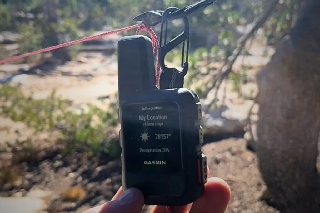 garmin-inreach-mini-2-weather-report Garmin inReach Mini satellite communicator held in hand, its monochrome screen showing an on-demand weather report with temperature and precipitation at a backcountry campsite.