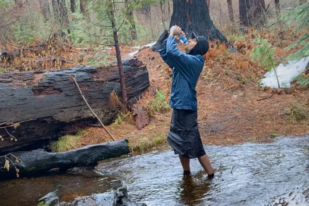 katadyn-befree-water-filter-drinking-in-stream Hiker standing in a shallow creek during rain, drinking from a filtered water bottle in a forest setting.