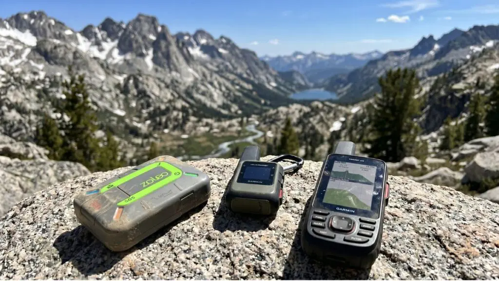 satellite-communication-devices-for-backpacking Three satellite communicators for backpacking — a green ZOLEO, a Garmin inReach Mini, and a Garmin GPSMAP — resting on granite above an alpine valley.