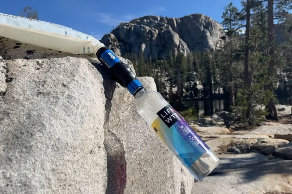 sawyer-squeeze-water-filter-gravity-setup Lifewtr bottle fitted with a Sawyer squeeze filter, propped on a granite boulder with a mountain peak and pine forest in the background.