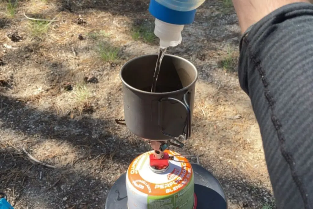 toaks-550-titanium-pot-backpacking-cook-kit Backpacker pouring filtered water into a small titanium pot on a canister stove, sunlit dirt clearing in the background.