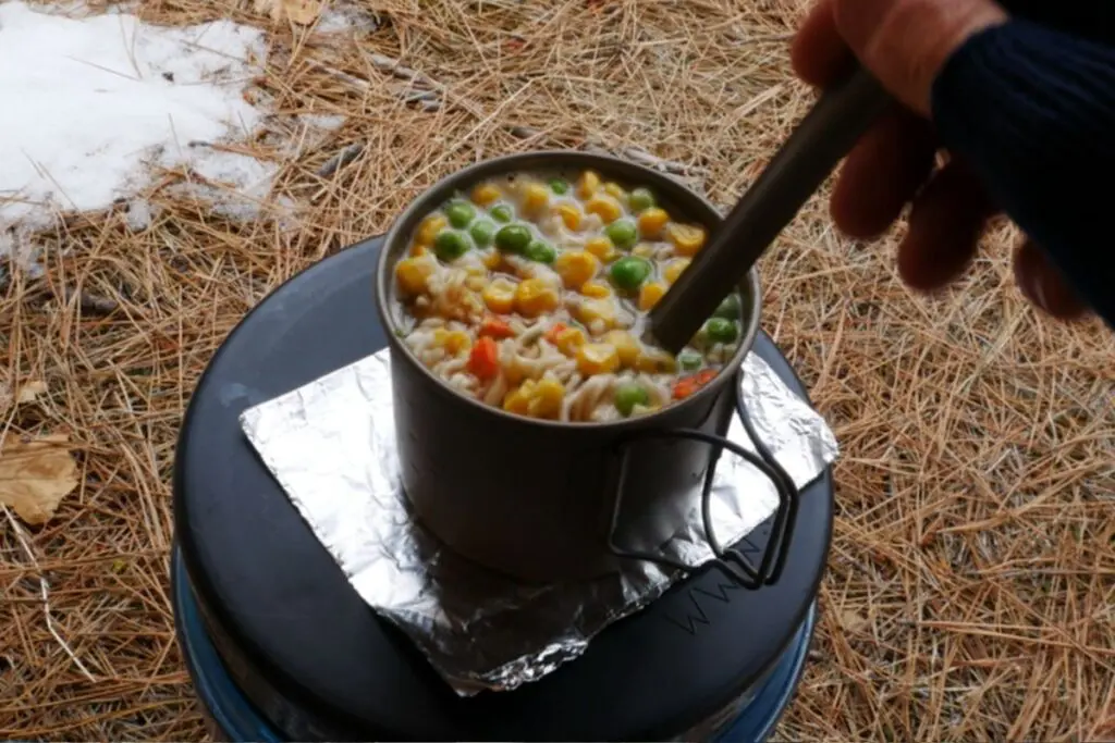 toaks-550-titanium-pot-backpacking-cook-ramen Ramen with corn and peas cooking in a titanium mug-pot resting on an inverted fuel canister with a foil windscreen.