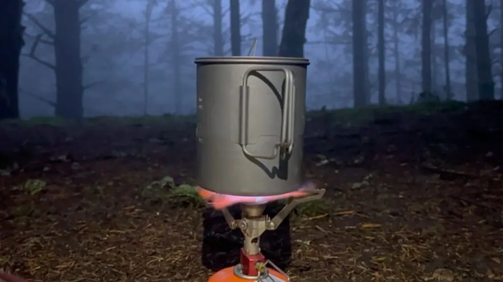 toaks-titanium-pot-backpacking-forest Titanium backpacking pot with a folding handle sitting on a canister stove, flames lit, in a misty evening forest.
