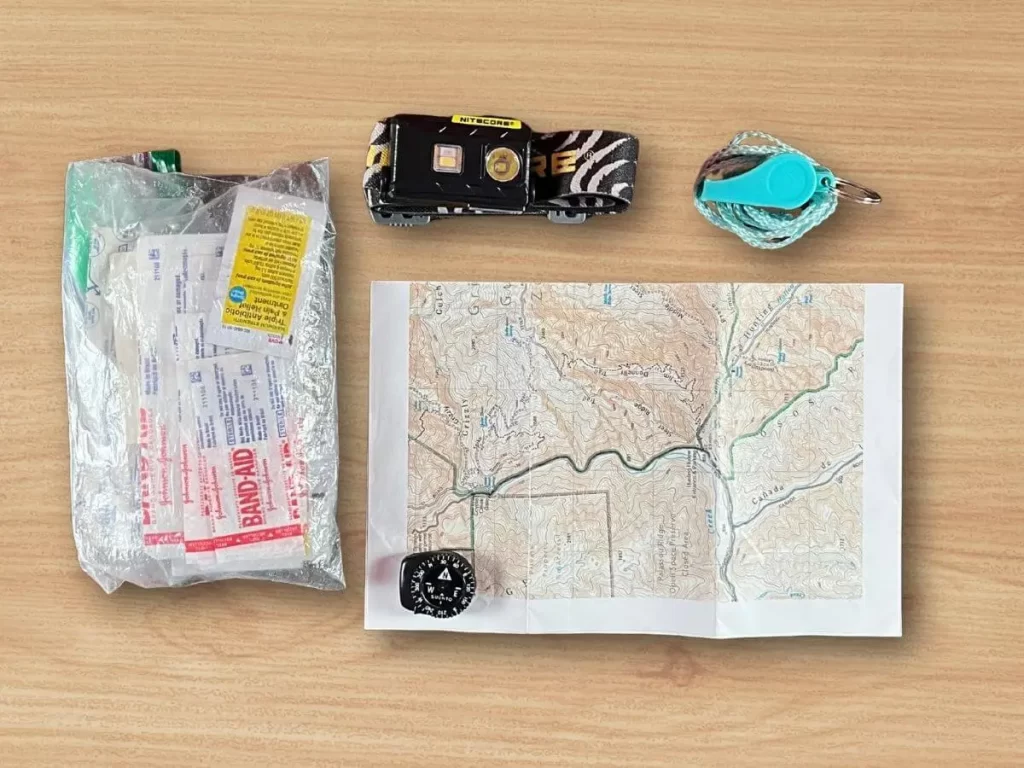 backpacking-navigation-first-aid-essentials Safety and navigation gear for backpacking, including a paper map, compass, first aid kit, headlamp, and emergency whistle.