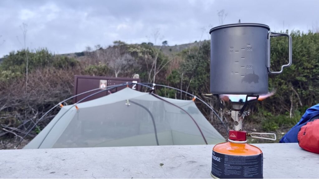 Backpacking stove with lit burner and pot on a canister at a coastal tent campsite.